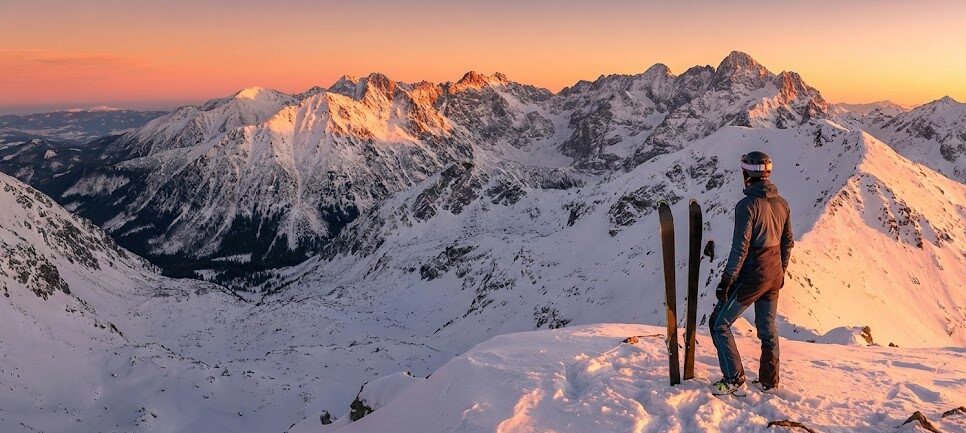 Panoramiczne ujęcie narciarza skiturowego na szczycie Grzesia o zachodzie słońca. Widoczne narty wbite w śnieg obok narciarza, który patrzy w stronę Tatr Wysokich.