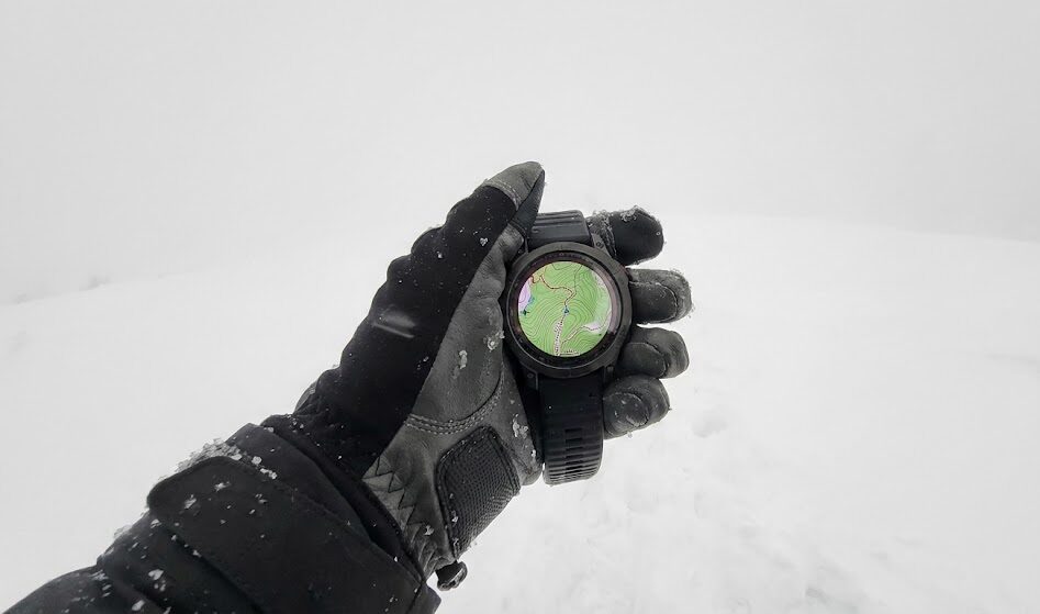 Zdjęcie wykonane z perspektywy pierwszej osoby (POV), przedstawiające dłoń w technicznej rękawiczce trzymającą nowoczesny smartwatch z kolorową mapą topograficzną.