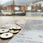 Zdjęcie paragonu fiskalnego z norweskiej restauracji leżącego na drewnianym stole obok norweskich monet i banknotów.