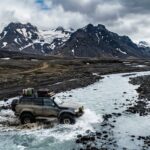 Przez środek płytkiej, lodowcowej rzeki powoli przeprawia się potężny, podniesiony samochód terenowy z napędem 4x4