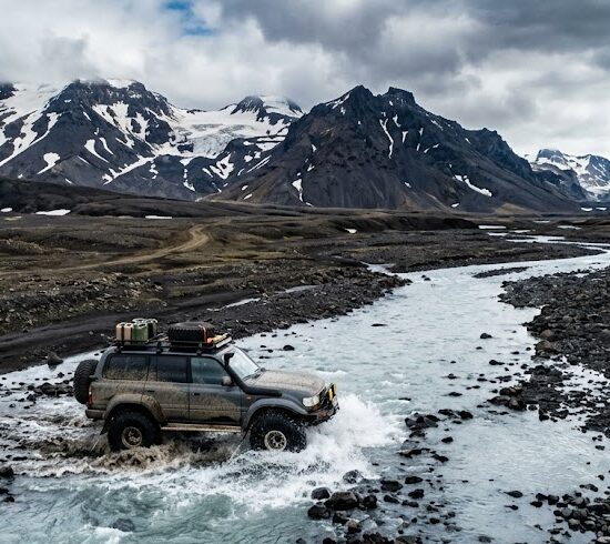 Przez środek płytkiej, lodowcowej rzeki powoli przeprawia się potężny, podniesiony samochód terenowy z napędem 4x4