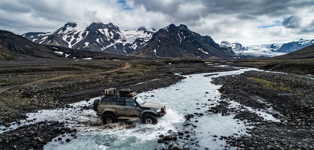Przez środek płytkiej, lodowcowej rzeki powoli przeprawia się potężny, podniesiony samochód terenowy z napędem 4x4