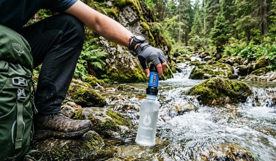 Turysta napełnia elastyczną butelkę (soft flask) z nakręconym na nią niebieskim, miniaturowym filtrem turystycznym